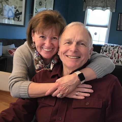 BMC South patient Peter Akeke, in casual dress and sitting in an armchair, smiling as he is embraced by his wife, Donna