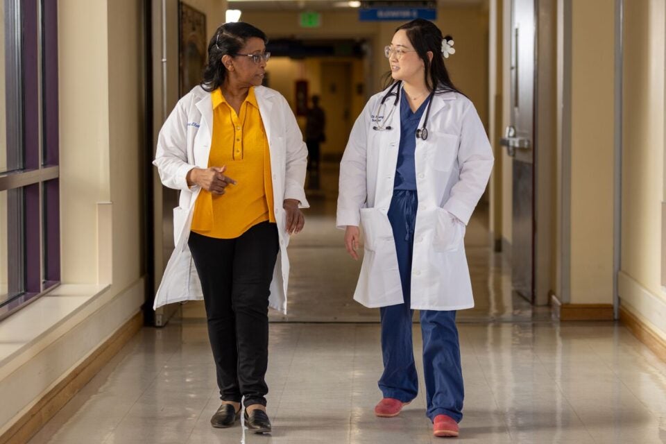 Two women wearing white lab coats, BMC Brighton pharmacist Salwa Elarabi (left) and resident physician Cathleen Huang (right), converse while walking down a hallway.