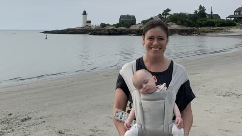 BMC Brighton epilepsy patient Lizzy stands on a New England beach while holding her sleeping baby in a front carrier (16x9 crop)