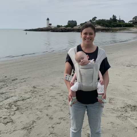 BMC Brighton epilepsy patient Lizzy stands on a New England beach while holding her sleeping baby in a front carrier