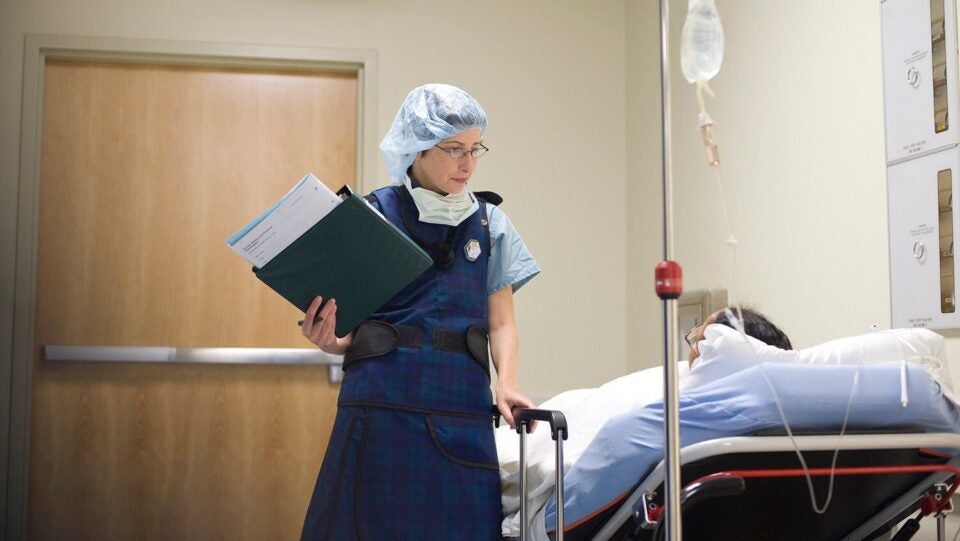 A female healthcare provider holding a clipboard of patient charts stands next to a patient lying on a hospital bed