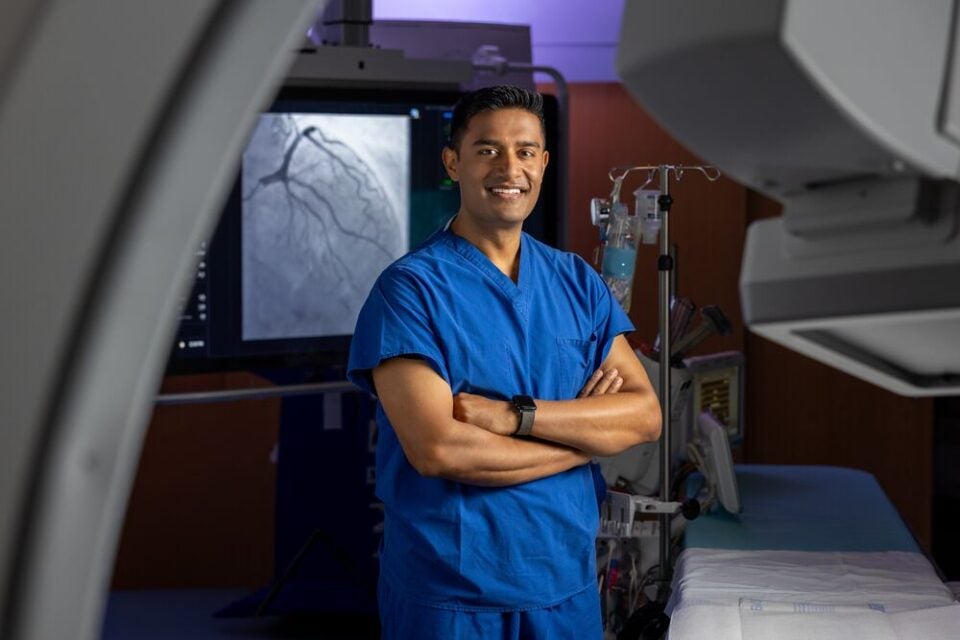 Sumon Roy, MD, FACC, FSCAI, interventional and structural cardiologist at BMC South, as well as BMC and BMC Brighton, stands smling with arms crossed in front of an operating table and a screen showing cardiac imaging