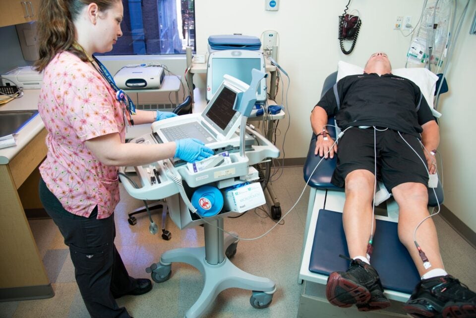 A middle-aged person in a black polo and black shorts lies on an exam table while hooked up to electrodes, as he is being evaluated for cardiac arrhythmia. A nurse in pink scrubs stands next to him looking at a computer monitor.