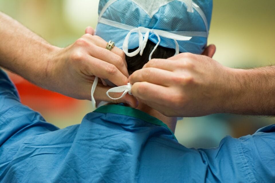 Close-up photo of the back and neck of a white, male surgeon wearing scrubs and a surgical cap as he ties a surgical mask before performing an operation