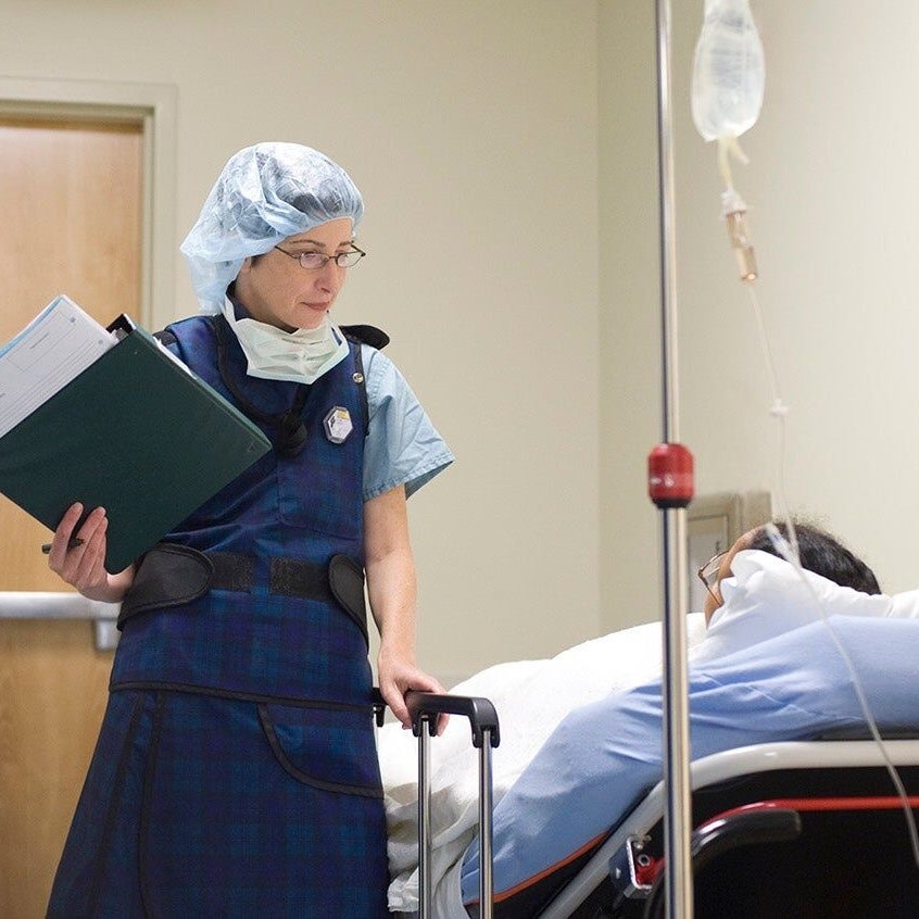 A female healthcare provider holding a clipboard of patient charts stands next to a patient lying on a hospital bed (1x1 crop)