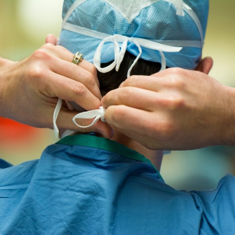 Close-up photo of the back and neck of a white, male surgeon wearing scrubs and a surgical cap as he ties a surgical mask before performing an operation (1x1 crop)