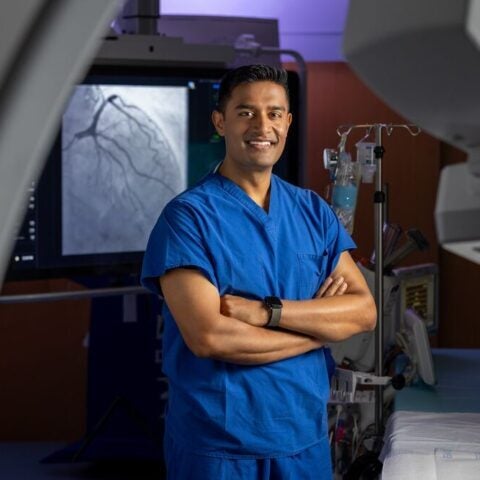 Sumon Roy, MD, FACC, FSCAI, interventional and structural cardiologist at BMC South, as well as BMC and BMC Brighton, stands smling with arms crossed in front of an operating table and a screen showing cardiac imaging
