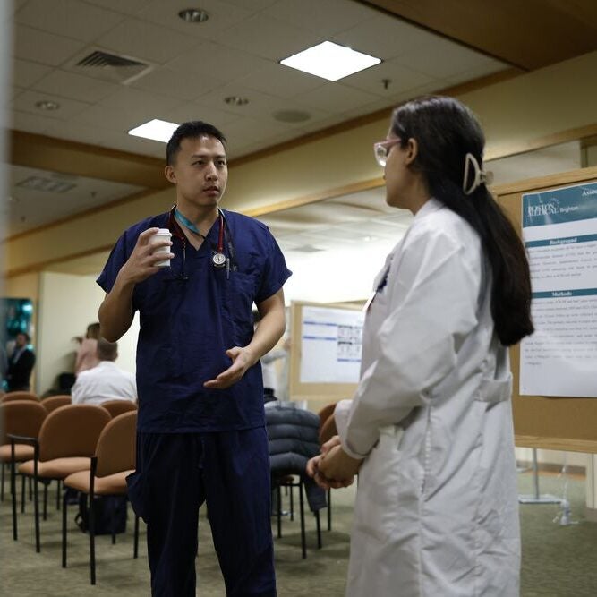 A person in navy scrubs holding a paper cup of coffee gestures while speaking with another person wearing a white lab coat, as they stand in a classroom in front of a clinical research poster about cardiovascular outcomes