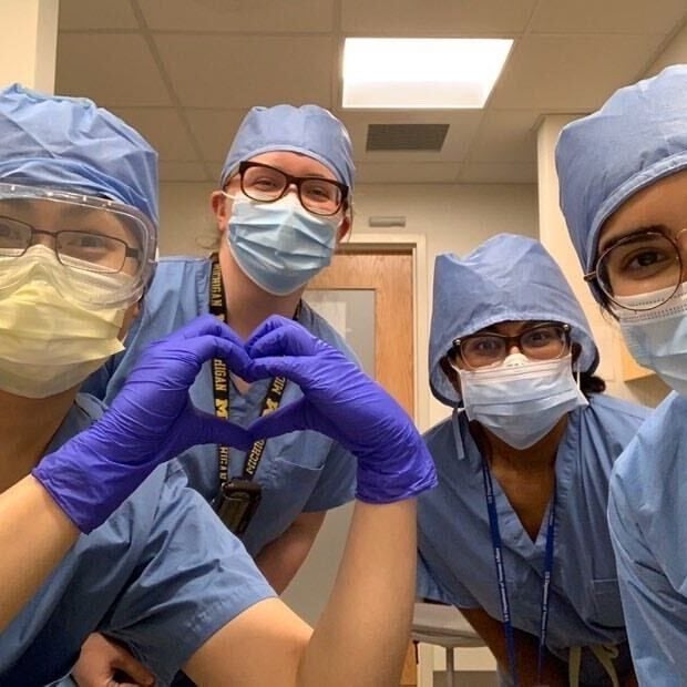 An internal medicine resident wearing full surgical scrubs, cap, and mask, forms her hands into the shape of a heart as three others stand behind her