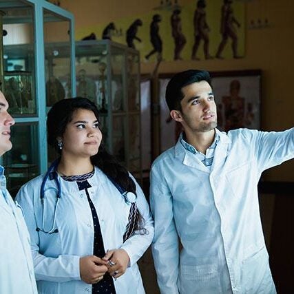 Young adults in lab coats observing medical images posted on a backlit wall. The person on the right is pointing to one of the images