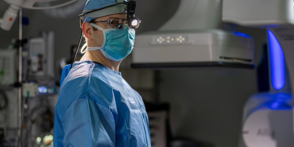 A man in surgical scrubs, mask, and head lamp stands in front of machinery in an operating theatre