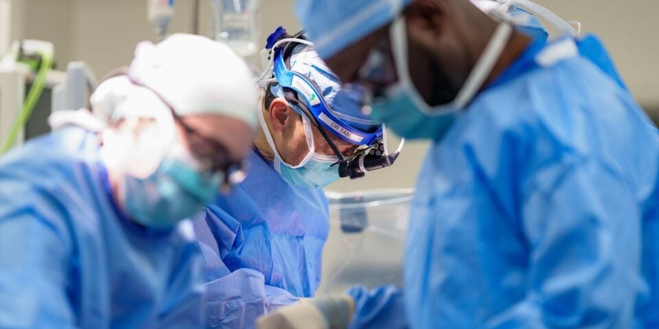 A group of three fully gowned and masked surgeons leans over a patient (not shown) on an operating table
